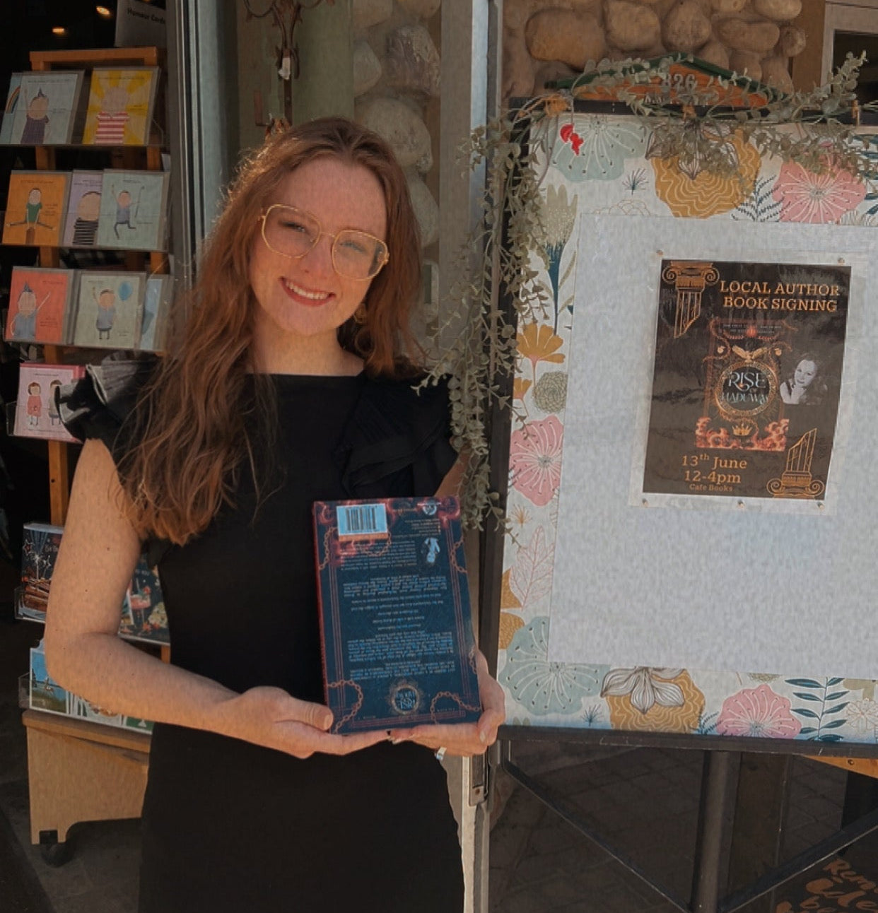 A woman in a black dress stands in a storefront doorway holding a blue book or booklet. A floral display board and shop interior are visible behind her.
